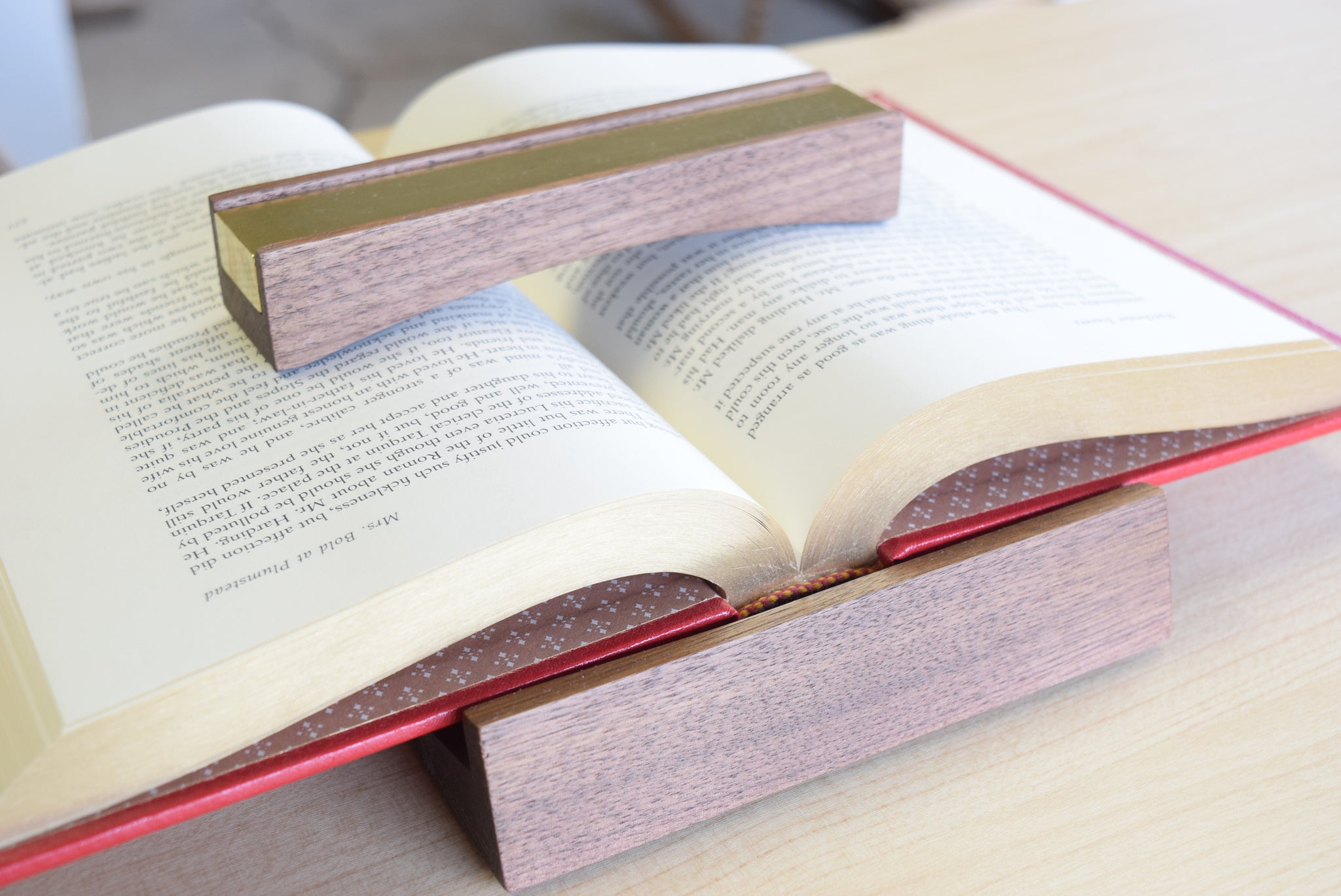 A walnut Bookfellow book rest shown holding the pages down of a book while simultaneously propping it up on a desk.