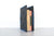 A black felt bookshelf or book holder with blue trim and four black buttons, holding two books, photographed on a light gray surface against a white brick wall.
