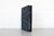 A dark gray Bookwarden slipcase shown stood up with a book in it, set against a white brick wall on a gray tabletop.