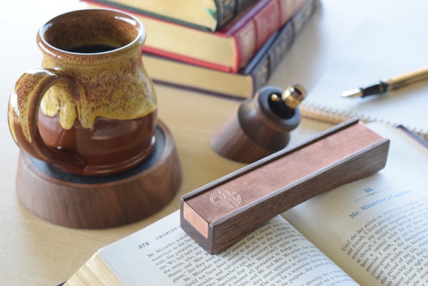 A wooden book weight and pen stand set on an open book, with a ceramic mug, books, and a pen in a softly lit desk scene.