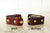 Two leather Nomad Book Weights displayed side by side on a marble surface against a white brick wall. The left Nomad shows the snap closure style, the right Nomad shows the button style closure style.
