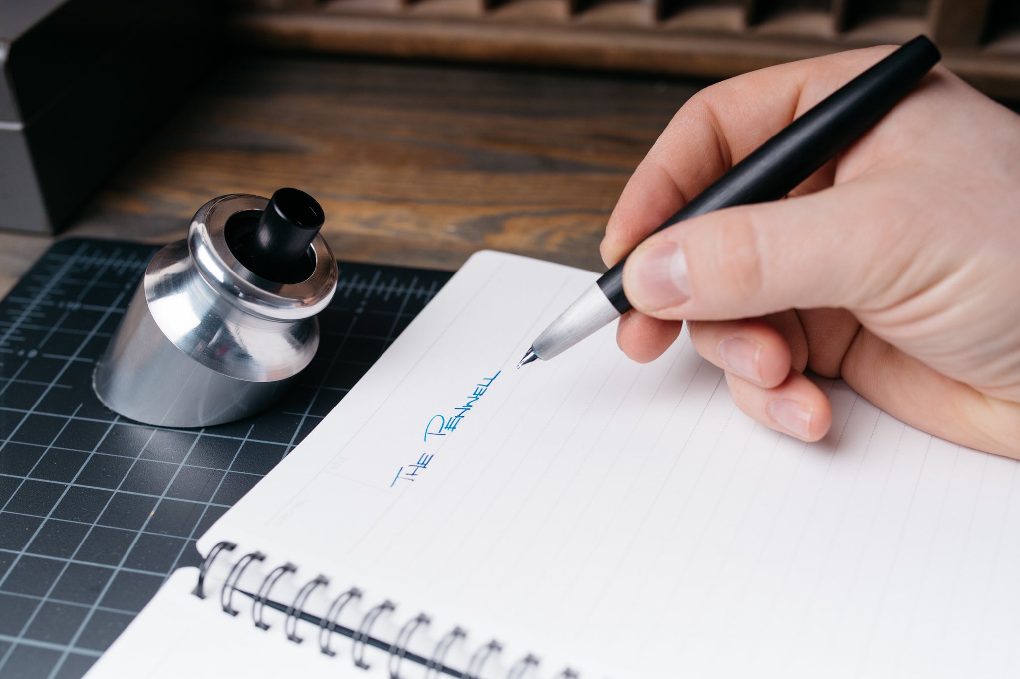 A close-up of a hand holding a black pen and writing in a white spiral notebook. A silver Penwell sits on a dark grid cutting mat in the background on a wooden desk.
