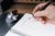 A close-up of a hand holding a black pen and writing in a white spiral notebook. A silver Penwell sits on a dark grid cutting mat in the background on a wooden desk.