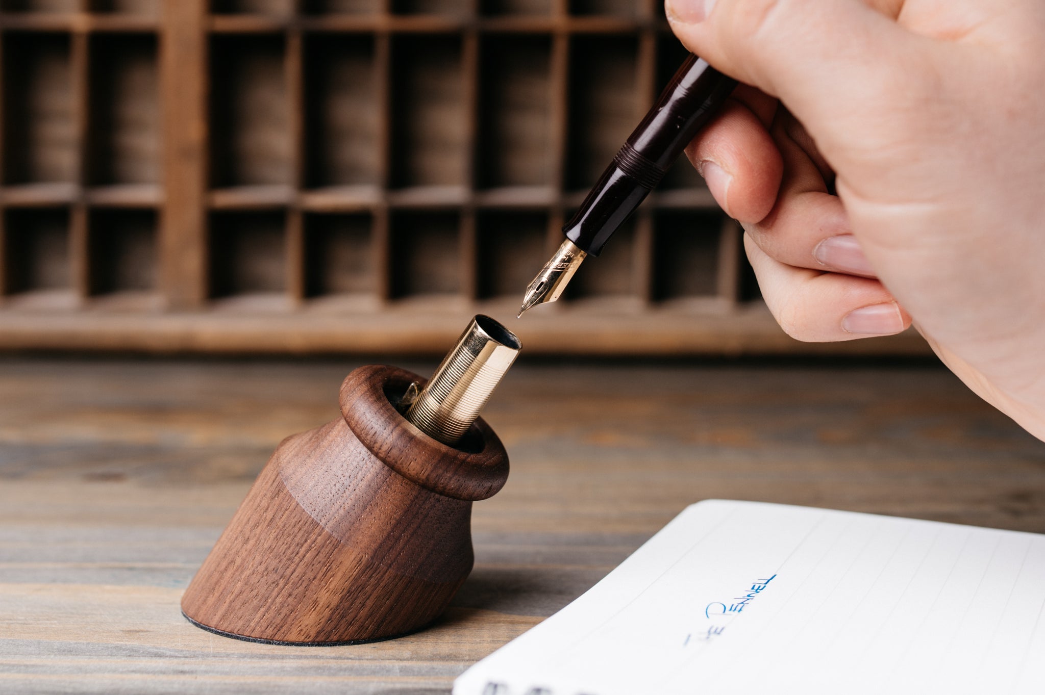 A walnut Penwell in use with a brown and gold fountain pen next to a notebook on a wood desk.
