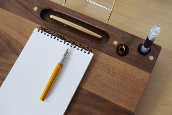 A portable desk with a walnut wood top, featuring a notepad, a yellow pen, and a penwell integrated into the surface. A brass door on the side reveals a pencil nook, and there is a clear acrylic page holder attached to the side of the desk.
