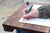 Someone writing in a notebook with a fountain pen on a brown wooden lap desk