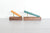 Two wooden pen stands on a light gray surface against a white brick wall. Each stand holds a slim pen resting diagonally: one pen is warm tan/orange wood with a black tip, and the other is teal-green wood with a gold tip.