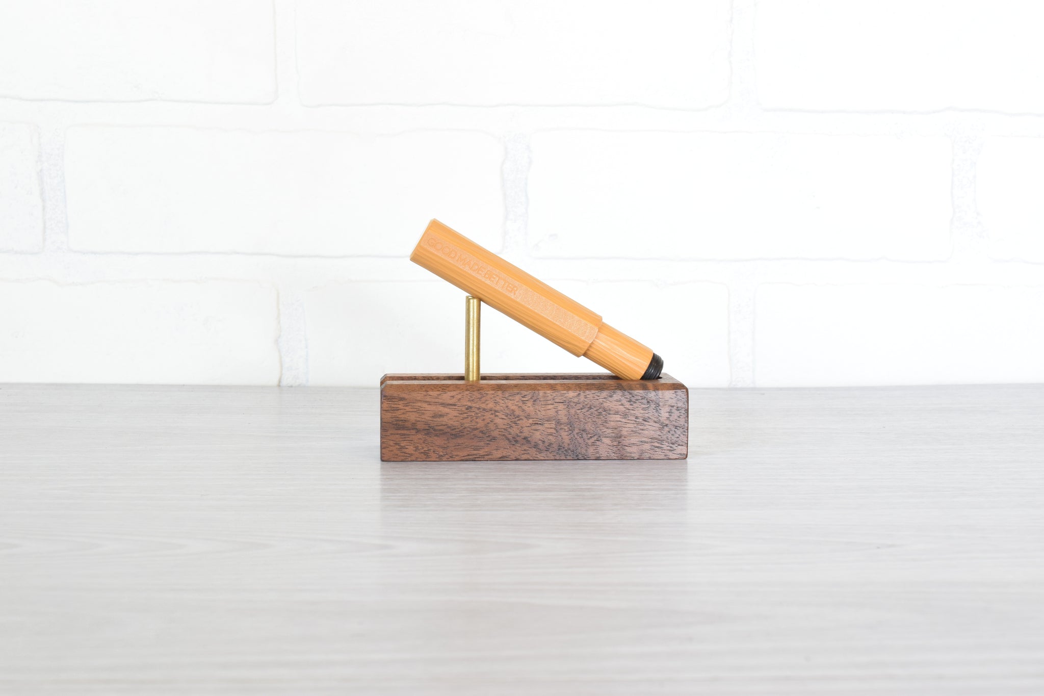 A mustard colored pen resting on a small brass stand atop a rectangular dark wood block, set against a white brick wall and light gray floor.