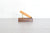 A mustard colored pen resting on a small brass stand atop a rectangular dark wood block, set against a white brick wall and light gray floor.
