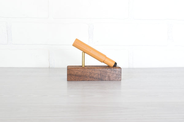 A mustard colored pen resting on a small brass stand atop a rectangular dark wood block, set against a white brick wall and light gray floor.