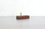 A wooden pen stand with upright brass supports, set on a light gray wooden tabletop against a white brick wall background.