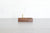 A rectangular dark wood pen stand with vertical brass rods, set on a light gray wood surface against a white brick wall background.