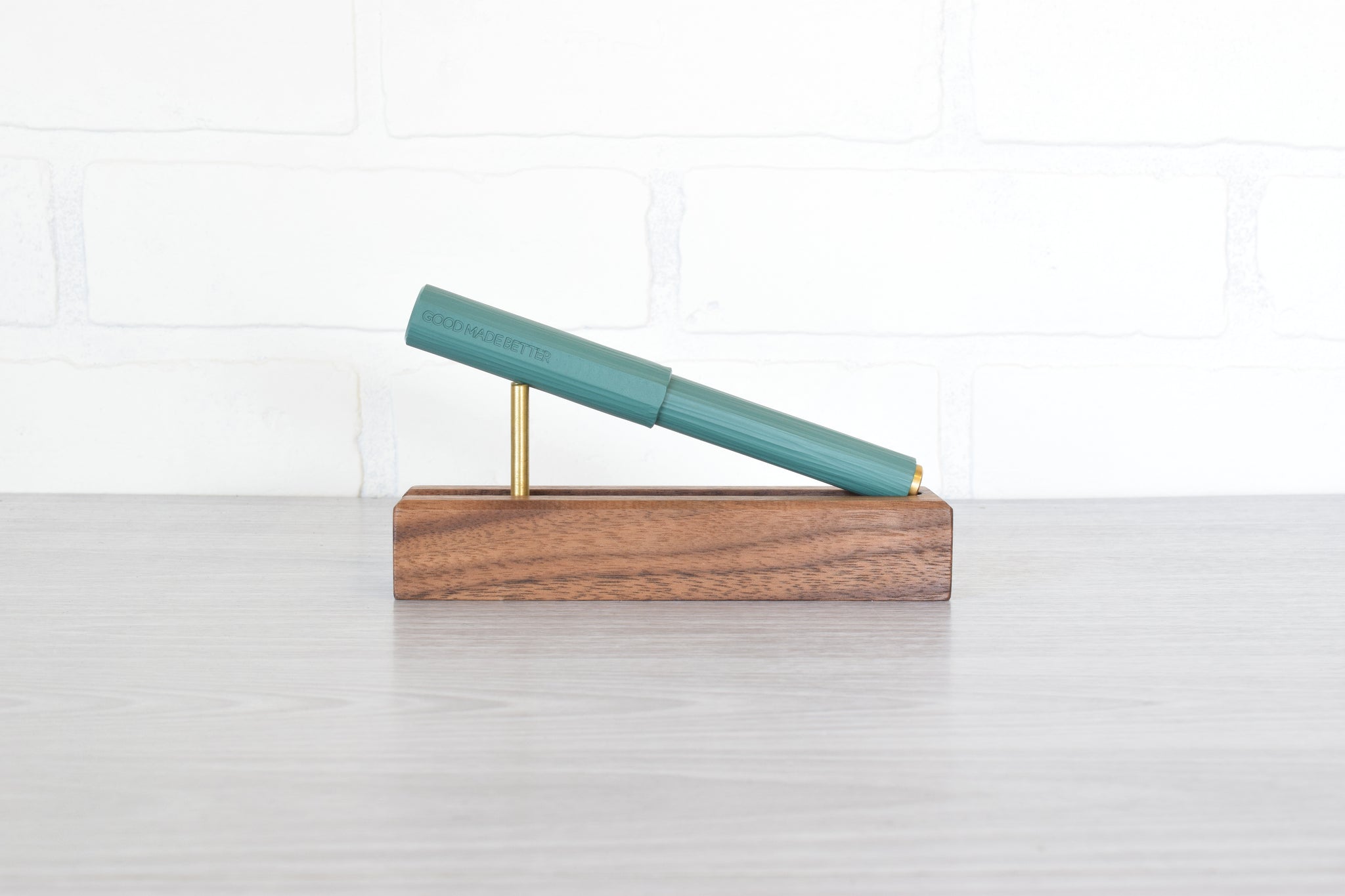 A teal green pen resting diagonally on a rectangular wooden display stand with brass metal support rods, set on a light gray tabletop against a white brick wall background.