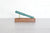 A teal green pen resting diagonally on a rectangular wooden display stand with brass metal support rods, set on a light gray tabletop against a white brick wall background.