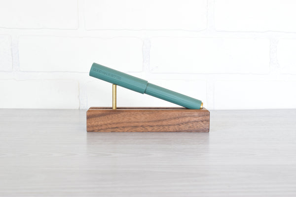 A teal green pen resting diagonally on a rectangular wooden display stand with brass metal support rods, set on a light gray tabletop against a white brick wall background.