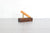A modern desk accessory featuring a dark brown wooden block base with a brass metal support holding a gold hued pen, set against a white brick wall and light gray tabletop.