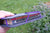 A hand holding a gray felt book holder with purple wire loops, containing books outdoors on a grassy lawn.