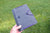 A hand holding a gray felt book holder with purple elastic bands and black buttons, set against a background of green grass.