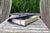 A dark gray felt book pouch with a purple elastic band, holding books on a weathered wooden table with tall green grass in the background.