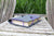 A dark gray felt book cover or journal sleeve with purple elastic bands holding a beige book, placed on a weathered wooden chair outdoors with tall green grass in the background.