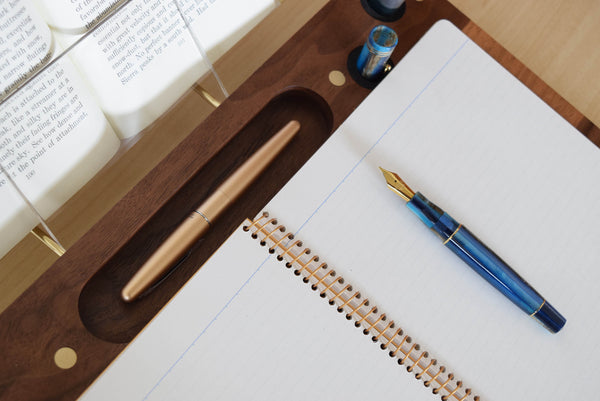 View from above a brown wood lap desk with a notebook, two fountain pens, and a book held open