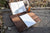Brown lap desk with notebook sitting on top, Two fountain pens and an open book being held, sitting on a stone outdoors
